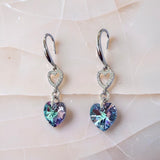 A pair of silver drop earrings with heart-shaped crystal charms, displayed on a light pink textured surface. The heart charms have an iridescent, color-shifting finish shifting between blue, green, and purple.
