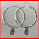 A pair of large silver hoop earrings with a Hamsa charm dangle.